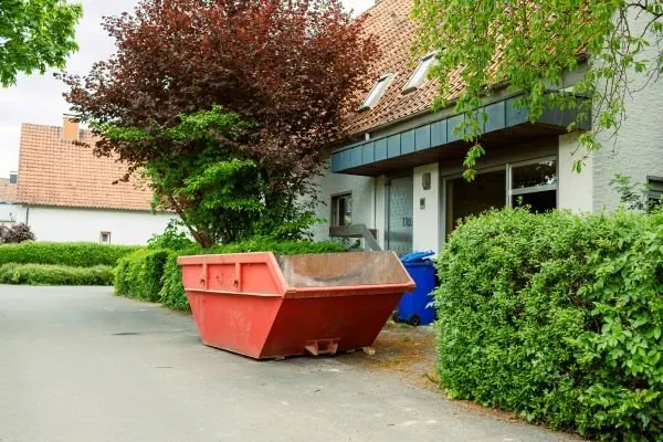 Small red open container on a residential driveway beside hedges and a brick house