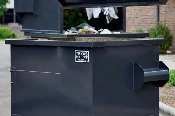 Dark front-load bin with its lid lifted, filled with household items and bags beside a brick wall