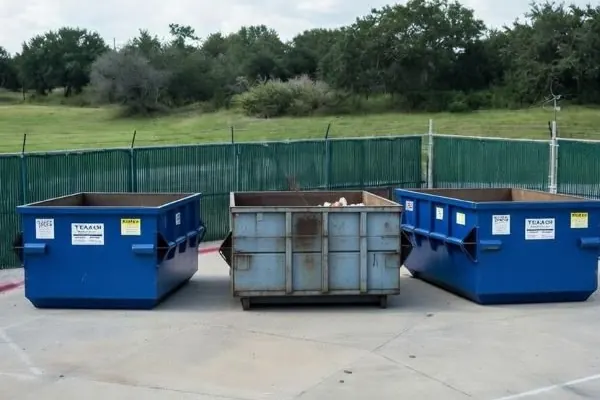 Three small open-top dumpsters lined up in a fenced lot beside a grassy field